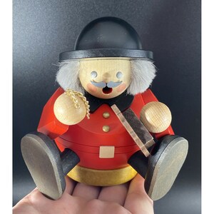 Könnte beinhalten: Eine Holzfigur eines Mannes, der einen roten Mantel, einen schwarzen Hut und einen weißen Bart trägt. Er hält eine goldene Kette in seiner rechten Hand und ein Holzobjekt in seiner linken Hand. Die Figur sitzt auf einem goldenen Sockel.