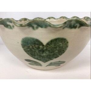 May include: A ceramic bowl with a cream-colored base and a scalloped rim featuring a green glaze. The exterior is decorated with a textured green heart shape positioned above two small green leaves, creating a simple and functional kitchen vessel.