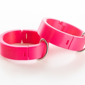 K&ouml;nnte beinhalten: Zwei verstellbare Plastikringe in leuchtendem Pink mit silbernen Metallverschl&uuml;ssen. Die Ringe sind zum Tragen am Handgelenk oder Kn&ouml;chel gedacht.