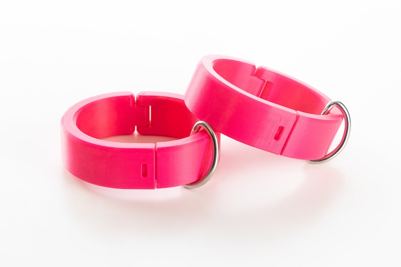 K&ouml;nnte beinhalten: Zwei verstellbare Plastikringe in leuchtendem Pink mit silbernen Metallverschl&uuml;ssen. Die Ringe sind zum Tragen am Handgelenk oder Kn&ouml;chel gedacht.