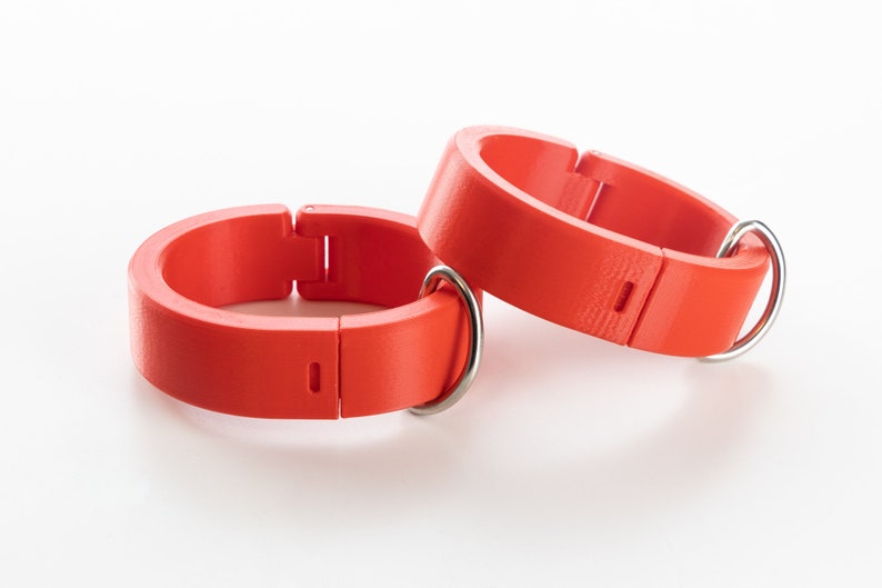 K&ouml;nnte beinhalten: Zwei leuchtend orangefarbene Kunststoffringe mit Metallverschl&uuml;ssen. Die Ringe sind verstellbar und haben eine glatte, leicht strukturierte Oberfl&auml;che.