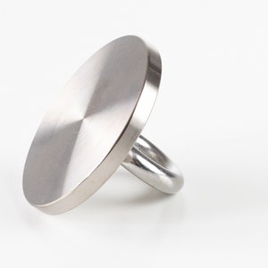 May include: A stainless steel ring with a large, circular face. The ring's face is a flat disc with a brushed metal finish. The band is a simple, rounded loop. The ring is set against a white background.
