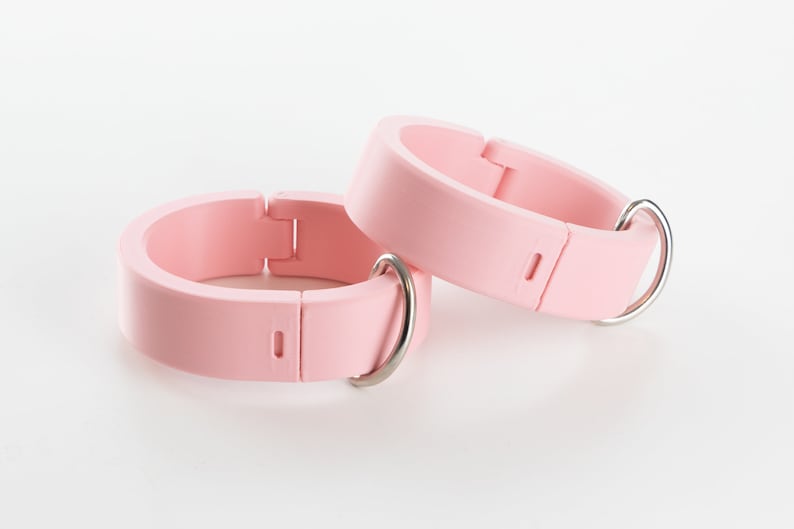 K&ouml;nnte beinhalten: Zwei rosafarbene Silikonringe mit silbernen Metallverschl&uuml;ssen. Die Ringe sind verstellbar und haben eine glatte, matte Oberfl&auml;che.