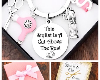 May include: A silver keyring with a pink hairdryer charm, a pair of silver scissors charm, a silver hairspray bottle charm, and a round silver charm with the text "This Stylist Is A Cut Above The Rest". The keyring is in a pink gift box with a white ribbon.