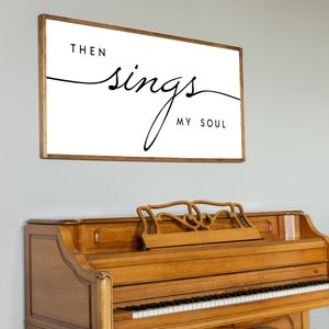 May include: A framed print with the text "Then sings my soul" in a cursive font. The print is hanging on a light grey wall above a wooden piano with a matching piano bench.