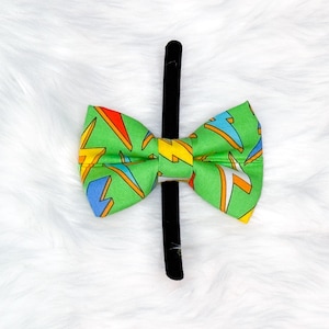 May include: A green bow tie with a repeating pattern of yellow, orange, and blue lightning bolts. The bow tie is attached to a black hair clip, set against a white, fluffy background.