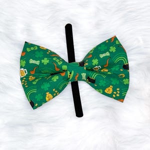 May include: A green St. Patrick's Day bow tie with a black center. The bow tie is patterned with gold coins, shamrocks, rainbows, beer mugs, and leprechaun hats. The bow tie is on a black headband.