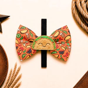 May include: An orange bow tie featuring a taco and food-themed pattern. A gold glitter taco with a smiling face is centered on a black band. The bow tie is surrounded by cowboy-themed accessories.