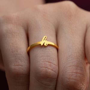 May include: A gold ring featuring the cursive letter "h". The ring is worn on a finger, with a blurred background. The ring's design is a simple band with the letter as a key feature. The ring is likely made of gold.