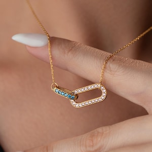 May include: A gold necklace with two interlocking oval pendants. One pendant is decorated with clear crystals, the other with blue crystals. The necklace is shown on a person's neck and hand.