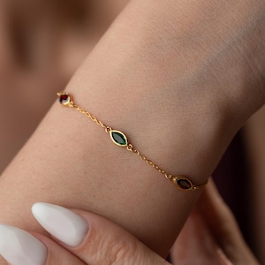 May include: A delicate gold bracelet featuring a series of colorful gemstones. The bracelet is adorned with a red, green, and purple gemstone. The bracelet is worn on a wrist.