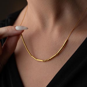 May include: A delicate gold necklace with a curved bar pendant. The pendant features small, evenly spaced cylindrical beads and a thin chain. The necklace is displayed against a dark background.