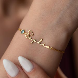Peut inclure: Un bracelet doré avec le nom "Sophia" écrit en cursive. Le bracelet est orné d'une petite pierre précieuse bleu clair et est porté au poignet. Bijou délicat et élégant.