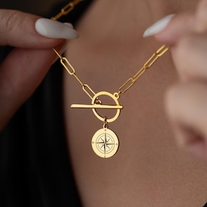 May include: Gold-colored necklace featuring a compass pendant. The necklace has a toggle clasp and a chain with rectangular links. The compass pendant is round with a detailed design. The necklace is being held up by a person.