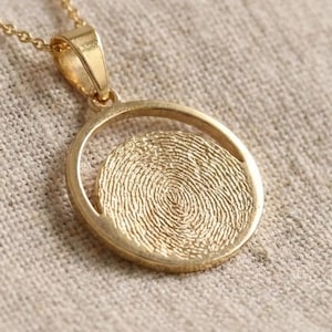 May include: A gold-colored pendant necklace featuring a circular pendant with a fingerprint design. The pendant hangs from a delicate gold chain. The background is a neutral-toned fabric.