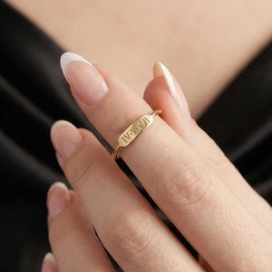 Può includere: Un anello in oro caratterizzato da una fascia sottile e una piccola placca rettangolare orizzontale incisa con i numeri romani IV-X-XI. Il metallo ha una finitura lucida e il design è centrato sul dito, con le lettere incise in scuro.