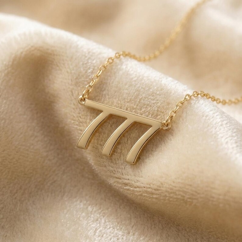 May include: A gold-colored necklace with a pendant featuring the number 777. The pendant is connected to a delicate chain. The necklace is displayed on a soft, beige fabric.