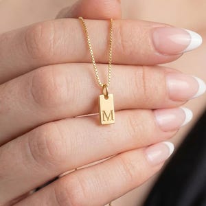 Op de afbeelding: Een goudkleurige ketting met een rechthoekige hanger met de letter "M". De hanger hangt aan een fijne ketting. De ketting wordt omhoog gehouden door een hand met verzorgde nagels.