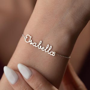 May include: A silver bracelet with the name "Isabella" in cursive script. The bracelet is attached to a delicate chain. The bracelet is worn on a wrist with white nail polish.