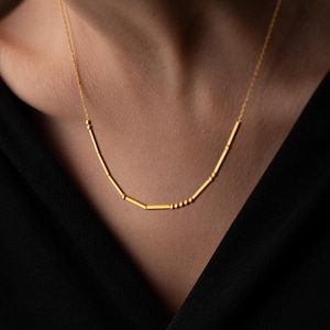 Könnte beinhalten: Eine zarte goldene Halskette mit linearem Design. Die Halskette hat eine Reihe kleiner, zylindrischer Perlen und eine feine Kette. Die Halskette wird vor einem dunklen Hintergrund getragen, der ihre helle, metallische Farbe hervorhebt. Die Halskette ist ein Schmuckstück.