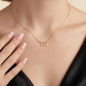 May include: A delicate gold-colored necklace featuring the number 777. The pendant is attached to a thin chain. The necklace is worn against a dark background.