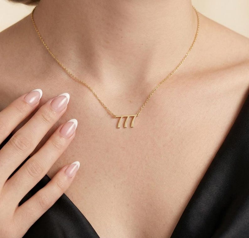 May include: A delicate gold-colored necklace featuring the number 777. The pendant is attached to a thin chain. The necklace is worn against a dark background.