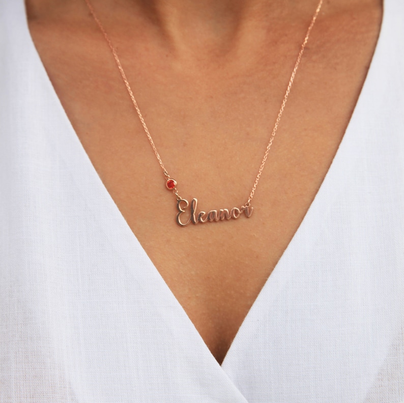 May include: A rose gold necklace with the name "Eleanor" spelled out in cursive script. A small red gemstone is attached to the chain above the name.