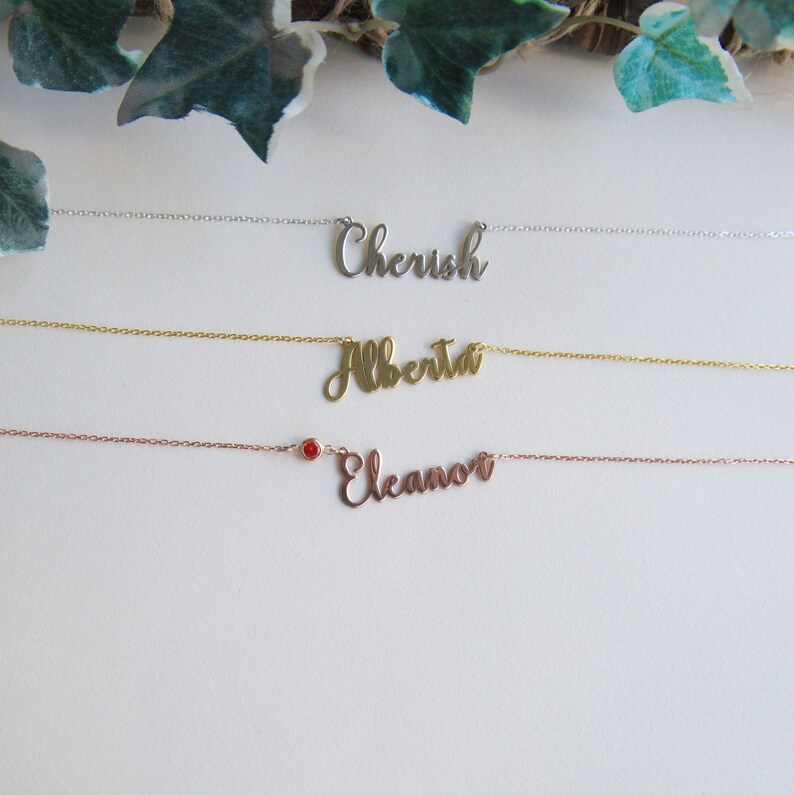 May include: Three delicate necklaces with personalized names. The top necklace is silver with the name "Cherish". The middle necklace is gold with the name "Alberta". The bottom necklace is rose gold with the name "Eleanor" and a small red gemstone.