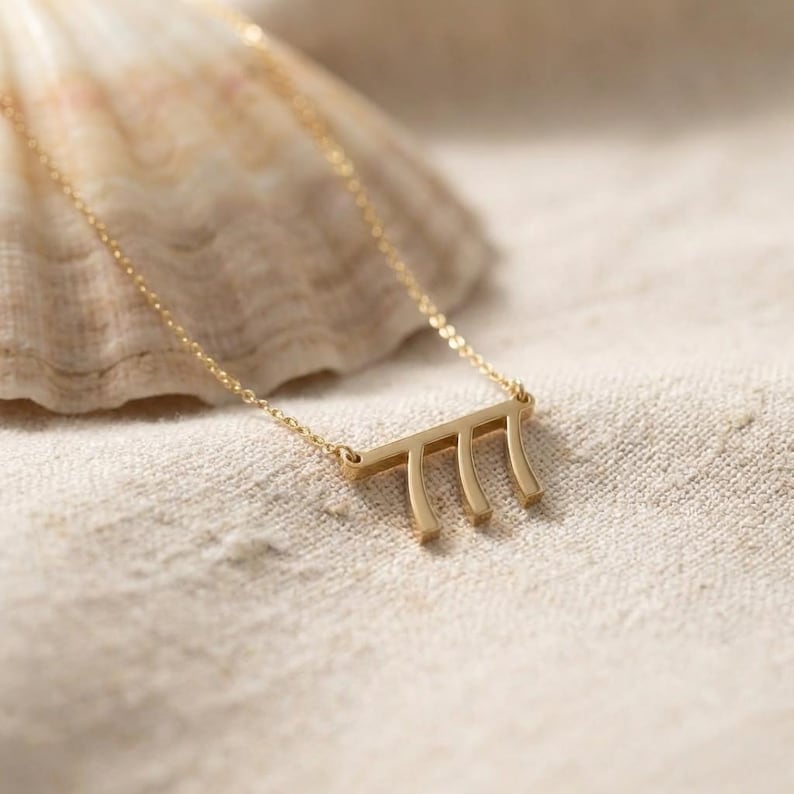 May include: A gold-colored necklace with a pendant featuring the number 777. The pendant is a horizontal bar with three vertical lines. The necklace is on a light-colored surface, with a seashell in the background.