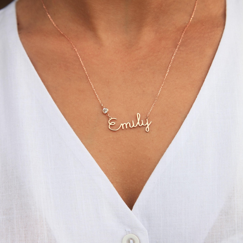 May include: A rose gold necklace with a small clear stone charm and a cursive script nameplate that reads 'Emily'