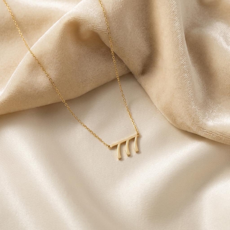May include: A delicate gold necklace featuring a pendant with the number 777. The pendant is a horizontal bar with three vertical lines. The necklace is displayed on a cream-colored, silky fabric.