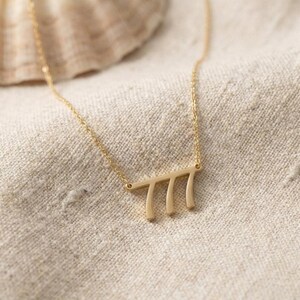 May include: A delicate gold-colored necklace with a pendant featuring the number 777. The pendant is a horizontal bar with three vertical lines. The necklace is displayed on a beige linen surface.
