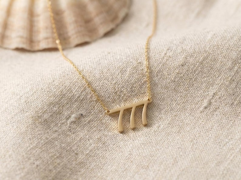 May include: A delicate gold-colored necklace with a pendant featuring the number 777. The pendant is a horizontal bar with three vertical lines. The necklace is displayed on a beige linen surface.