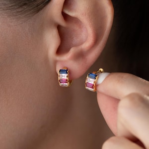 May include: Gold-coloured hoop earrings with a rectangular design. Each earring features a row of gemstones in blue, white, and red. The earrings are shown on an ear and being held by a hand.