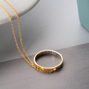 Könnte beinhalten: Goldfarbene Halskette mit einem kreisförmigen Anhänger, auf dessen äußerem Rand römische Ziffern eingraviert sind. Der Anhänger hängt an einer filigranen Kette. Die Halskette wird auf einer grauen Oberfläche präsentiert.