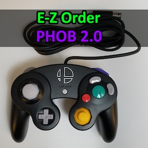Puede incluir: Un mando de GameCube negro con un botón blanco y un botón amarillo. El mando tiene un botón morado y verde en la parte superior y un cable negro conectado a él. El texto "E-Z Order PHOB 2.0" es visible en la imagen.