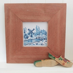 May include: A framed cross-stitch depicting a blue and white Dutch windmill scene with a house and a bridge. The frame is made of wood and has a reddish-brown stain. Two wooden clogs with green and red painted designs are in the foreground.