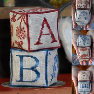 Può includere: Una pila di tre blocchi di tessuto ricamato, ognuno con una lettera e un design diversi. Il blocco superiore è rosso e bianco con la lettera 'A' e un design floreale. Il blocco intermedio è blu e bianco con la lettera 'B' e un design floreale. Il blocco inferiore è rosso e bianco con un design floreale.