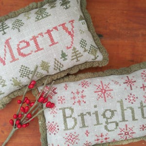 May include: Two rectangular Christmas pillows with cross-stitched text and designs. One pillow says "Merry" in red, the other says "Bright" in green. Both have green trim and tree and snowflake patterns.