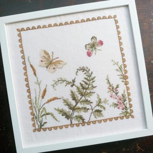 May include: A white framed embroidery featuring a butterfly, fern, and flowers in shades of green, brown, and pink. The embroidery is surrounded by a gold border.