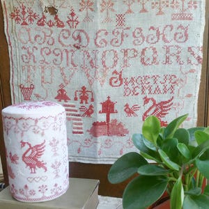 May include: A vintage needlework sampler with red cross-stitch letters and motifs, including birds and trees, hangs behind a cylindrical container with matching swan and floral designs. The container sits on a stack of old books, with a small house-shaped planter in the foreground.