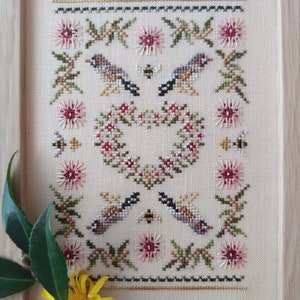 Puede incluir: Una bordadura de punto de cruz enmarcada que presenta una corona de flores en forma de corazón, dos pájaros y abejas. El diseño está hecho en tonos de rosa, verde, marrón y dorado.
