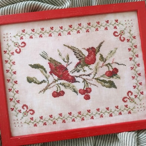 Pode incluir: Obra de arte em ponto cruz emoldurada em vermelho. A peça apresenta dois pássaros vermelhos empoleirados em um galho com cerejas. O design é cercado por uma borda decorativa com detalhes em vermelho e verde. A moldura é vermelha vibrante.