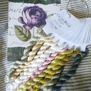 May include: A close-up of a collection of embroidery floss spools in various shades of green, yellow, pink, and white. The spools are arranged in a fan shape and labelled "CASCADE HOUSE Embroidery Stranded Silk Colour 7880".