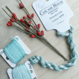 May include: Two spools of light blue embroidery floss, labelled 3761 and 1912, with a strand of the floss in a twisted braid. A white label with the text "CASCADE HOUSE EMBROIDERY Since 1994 Stranded Silk Colour 8395 7.5 Metres" is in the background.