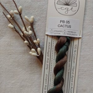 May include: A skein of stranded cotton embroidery floss in shades of green and brown. The floss is labelled "PB 05 CACTUS" and is 100% cotton, hand-dyed in Australia. The label also states that the floss is 10 metres pre-cut in 50 cm lengths.