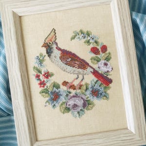 May include: A framed cross-stitch embroidery featuring a bird with a crest, brown and white plumage, and a red tail, positioned within a circular wreath of colorful flowers and berries on a light beige fabric background in a light wood frame.