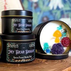 May include: Three black metal candle tins with white lids. Each tin has a white candle with a mushroom and flowers embedded in the wax. The candle tins have a green label with the text "MY DEAR DRAGON" and "Green Forest Herbs | Soy Wax Candle".