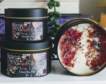 May include: Three black candle tins with gold lids. The candles are decorated with dried flowers and glitter. The text on the labels reads "You've Survived Now Live" and "Citrus Sea Mist".