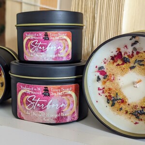 Three Starborn Candles in elegant black tins with gold accents, displayed on a white shelf. Each candle features a colorful label detailing the scent of lilac, nutmeg, and honeyed spice. The front candle is open, showcasing its luxurious wax topped with crushed rose petals and vibrant crystal chips, while the two closed tins highlight their beautiful design. The background includes wooden decor and plants, enhancing the cozy aesthetic.